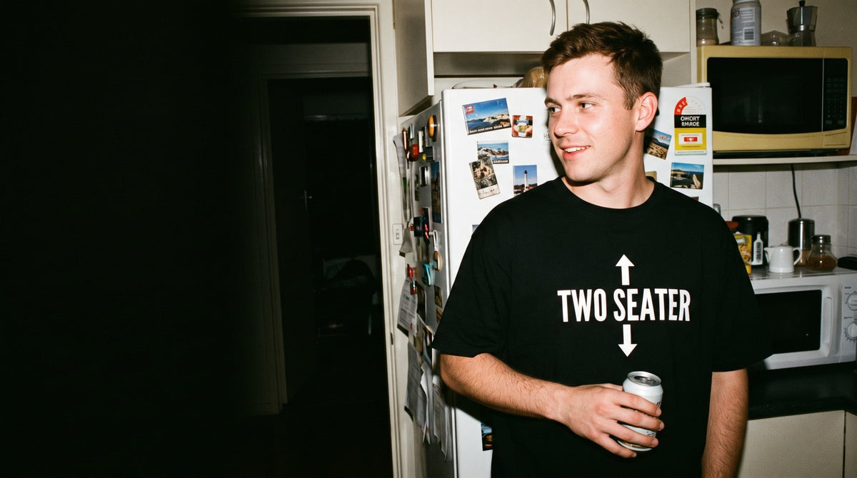 Guy in a black Two Seater tee holding a can in a share-house kitchen